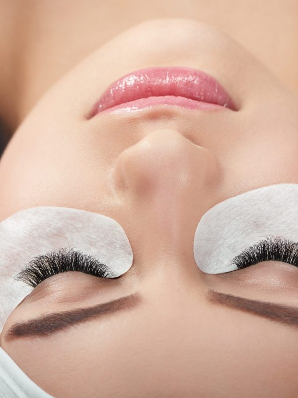 Photo comparison of woman's normal and fake enlarged lashes. Client lying on special coach wearing headbandage for lashmaking. Having black hair. Loving beaty procedures in salone. Pretty young face.